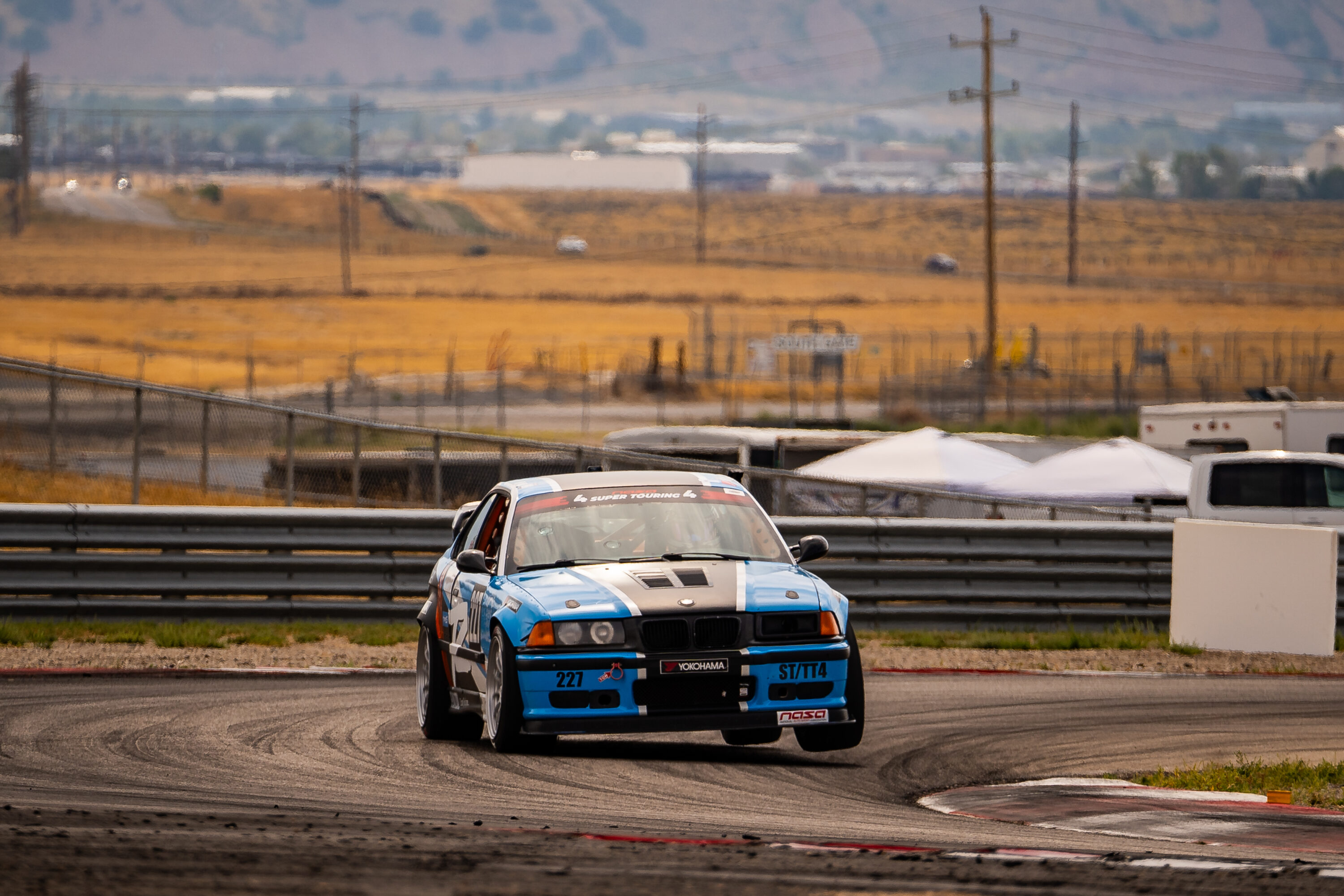 Blue and white BMW E36 race car #227 cornering on track at a NASA event, getting two wheels in the air.