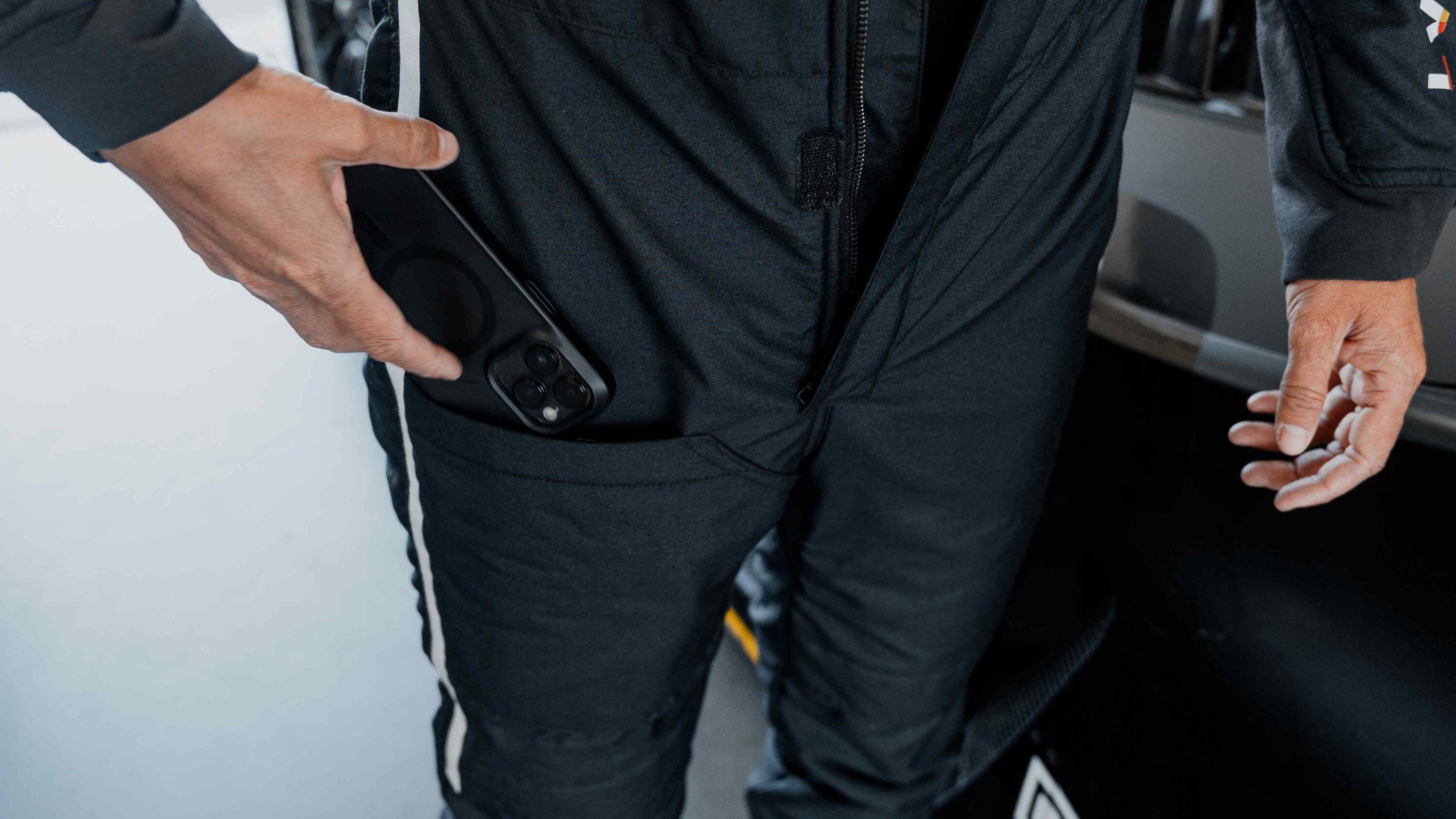 Close-up of the LVRY DRVR racing suit showing a driver sliding a phone into the built-in mobile pocket, highlighting functional storage and ergonomic design.