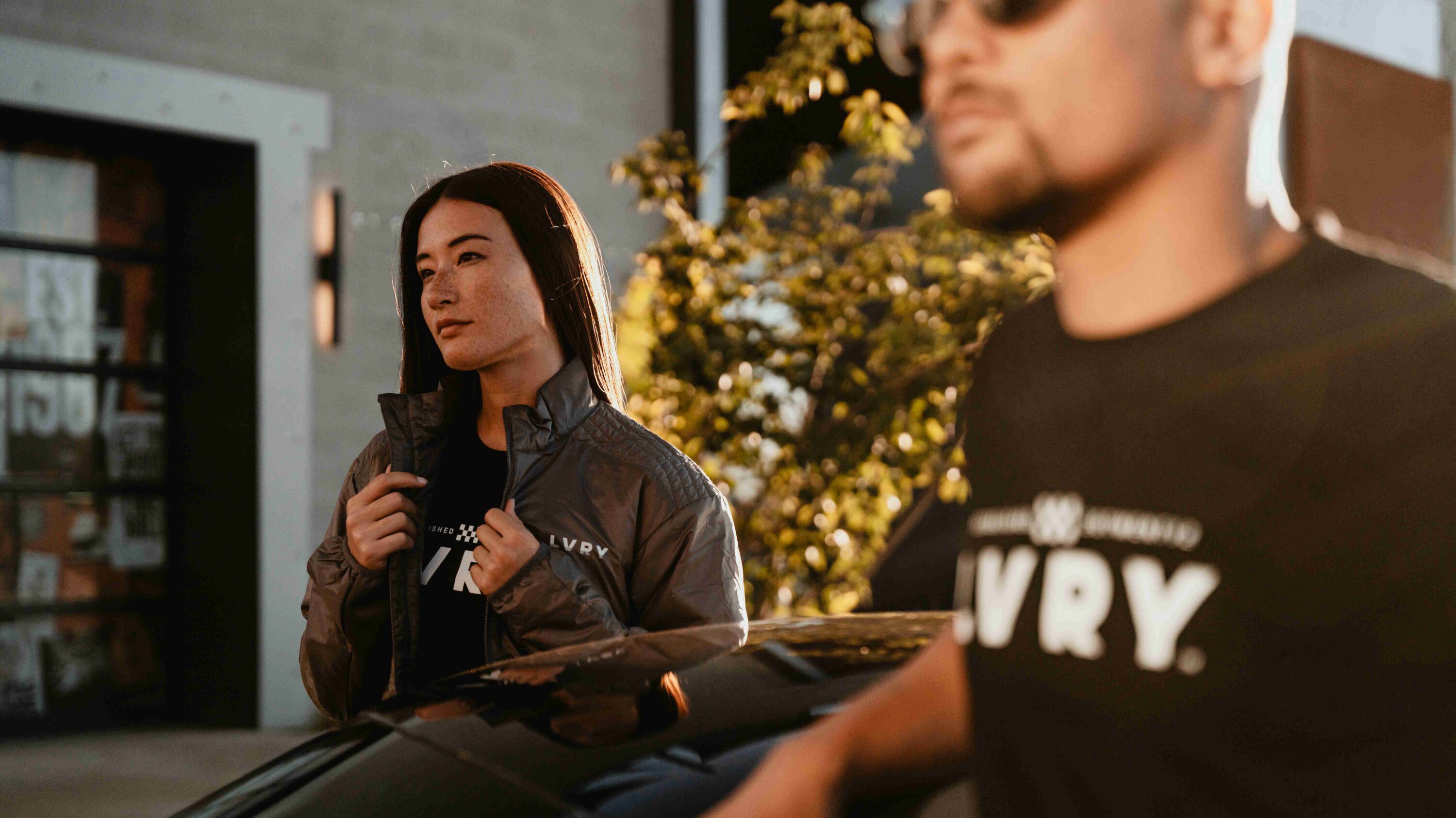 Driver wearing LVRY DRAFT Insulator lightweight jacket beside sports car at golden hour, showcasing race-inspired performance outerwear.