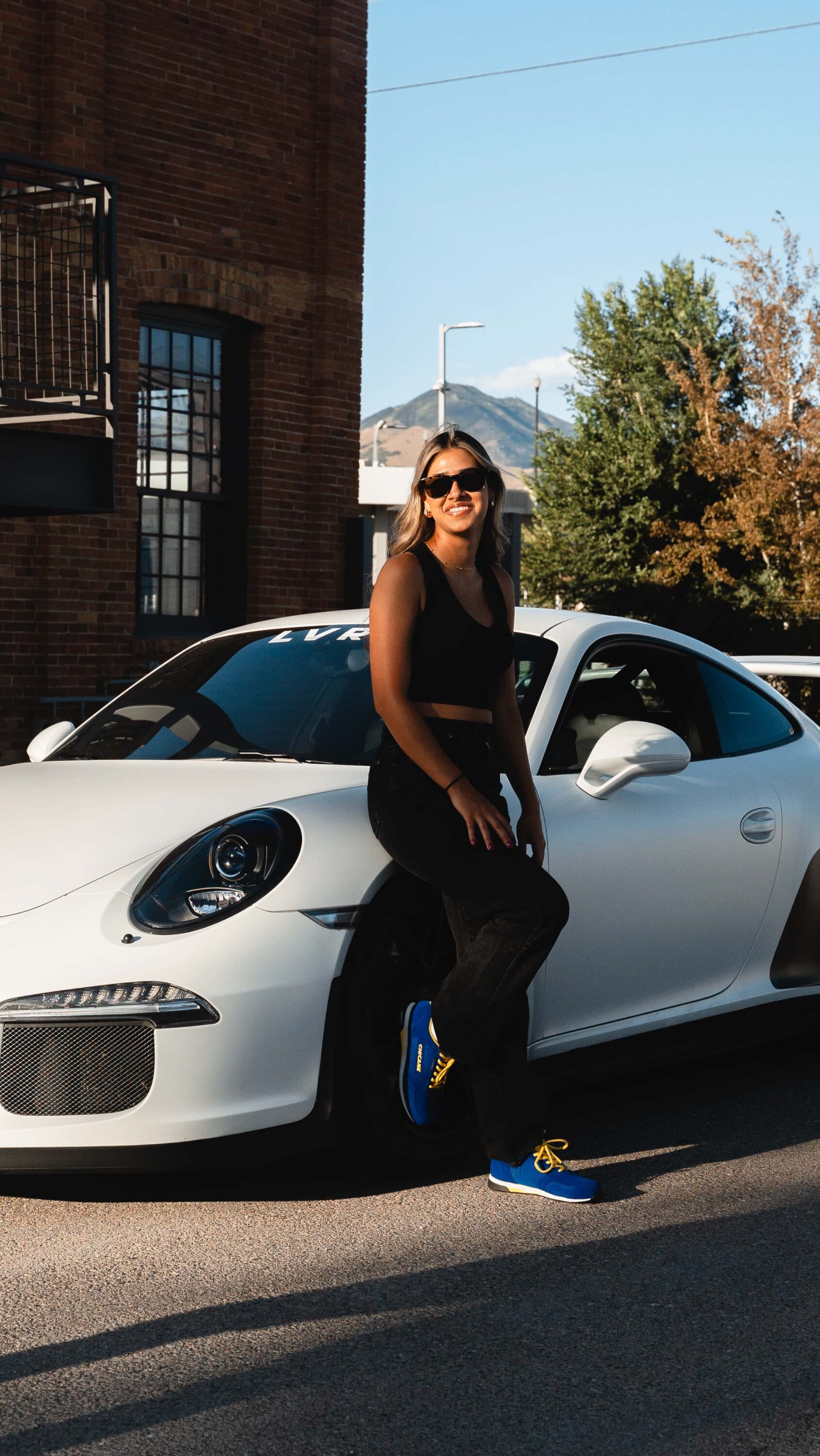 Woman leaning on white Porsche wearing blue Chicane Sebring women’s driving shoes in urban setting, highlighting motorsport lifestyle and fit.