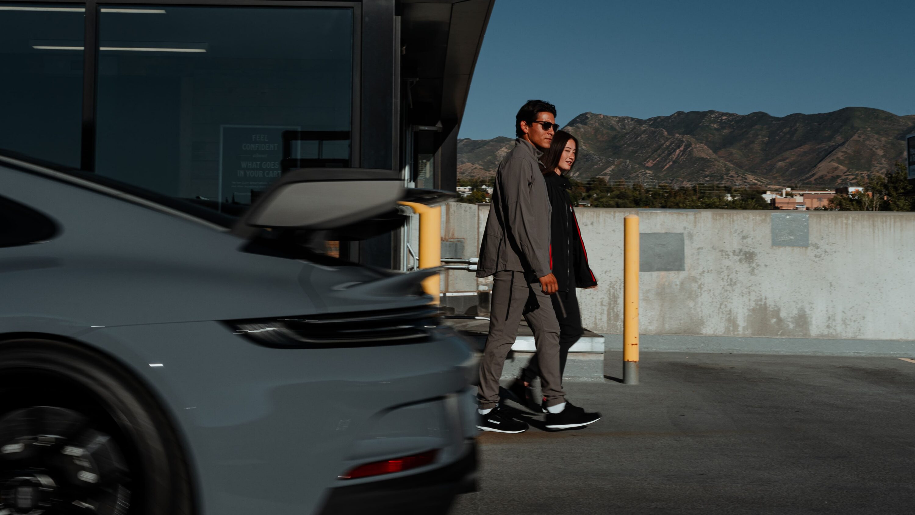Man and woman walking past a sports car in a parking structure wearing LVRY SECTR 1 technical pants designed for movement between track and street.