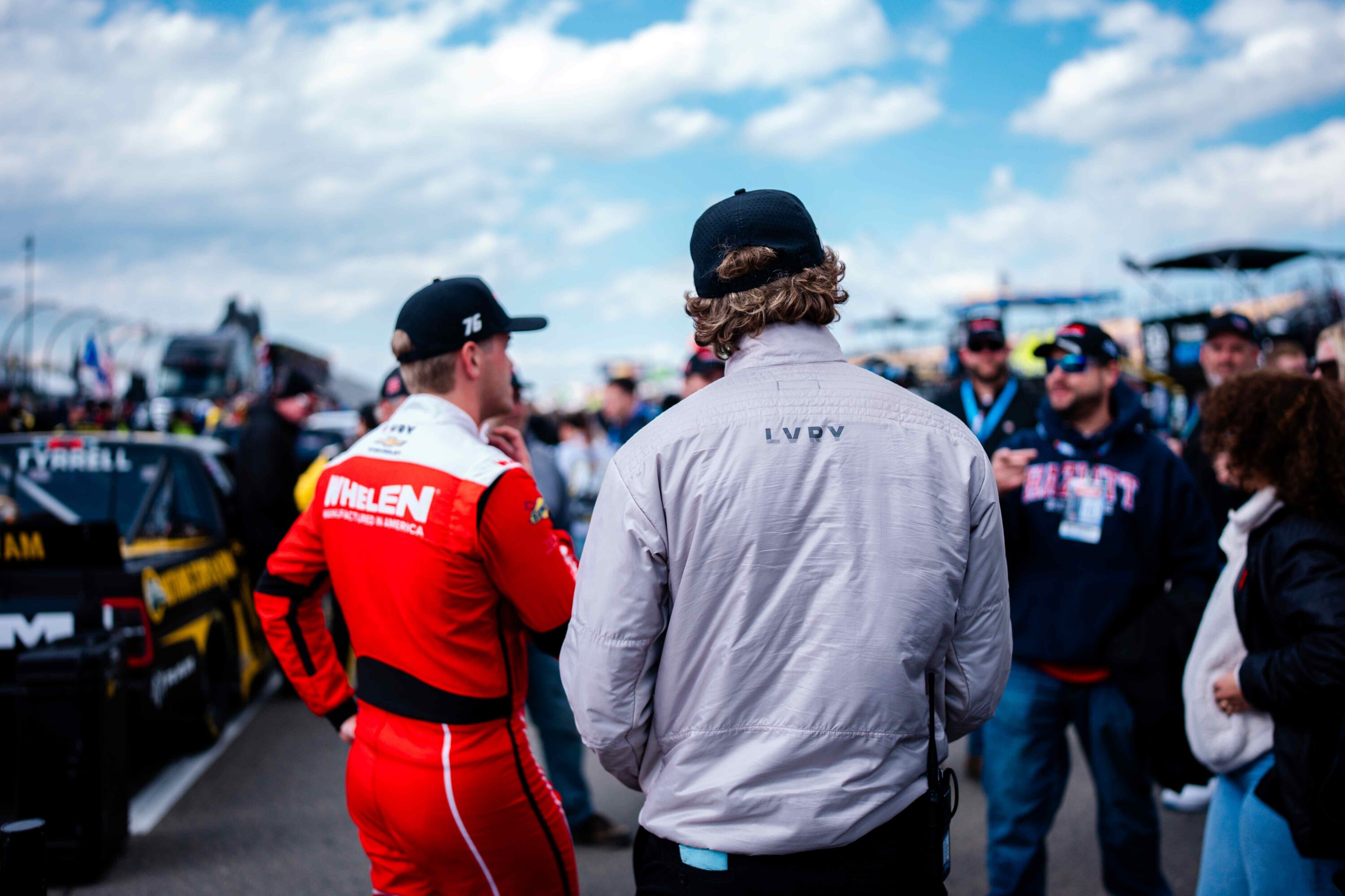 LVRY DRAFT ultra-light insulated jacket worn at Watkins Glen NASCAR race weekend, shown from behind in light gray with subtle LVRY branding, paired alongside a driver in a red Whelen racing suit on pit road.