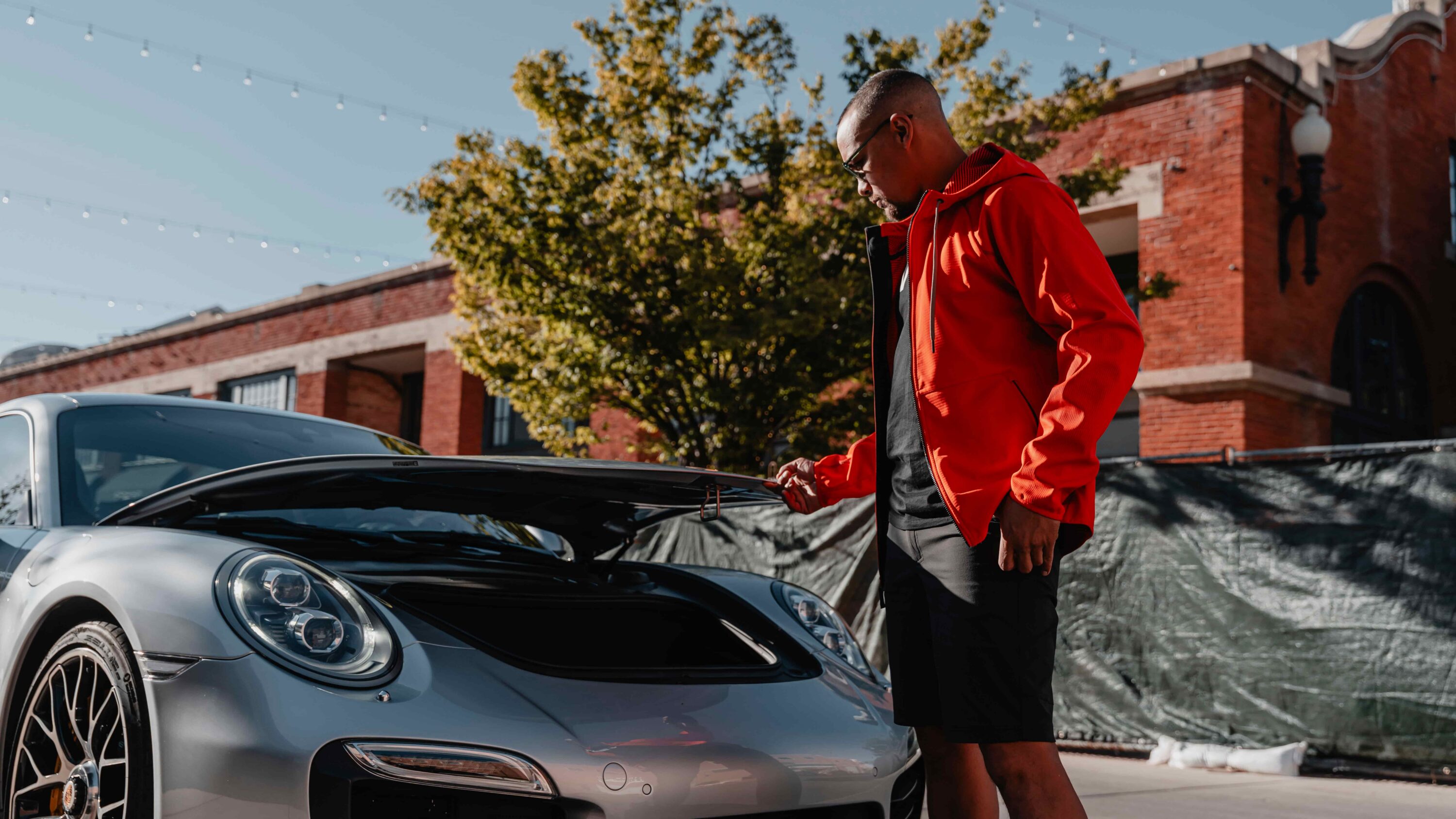 Man in a red LVRY PACE jacket and SPRINT shorts leaning over the hood of a Porsche on a warm sunny day, showing the nylon-spandex stretch system built for hot track days.