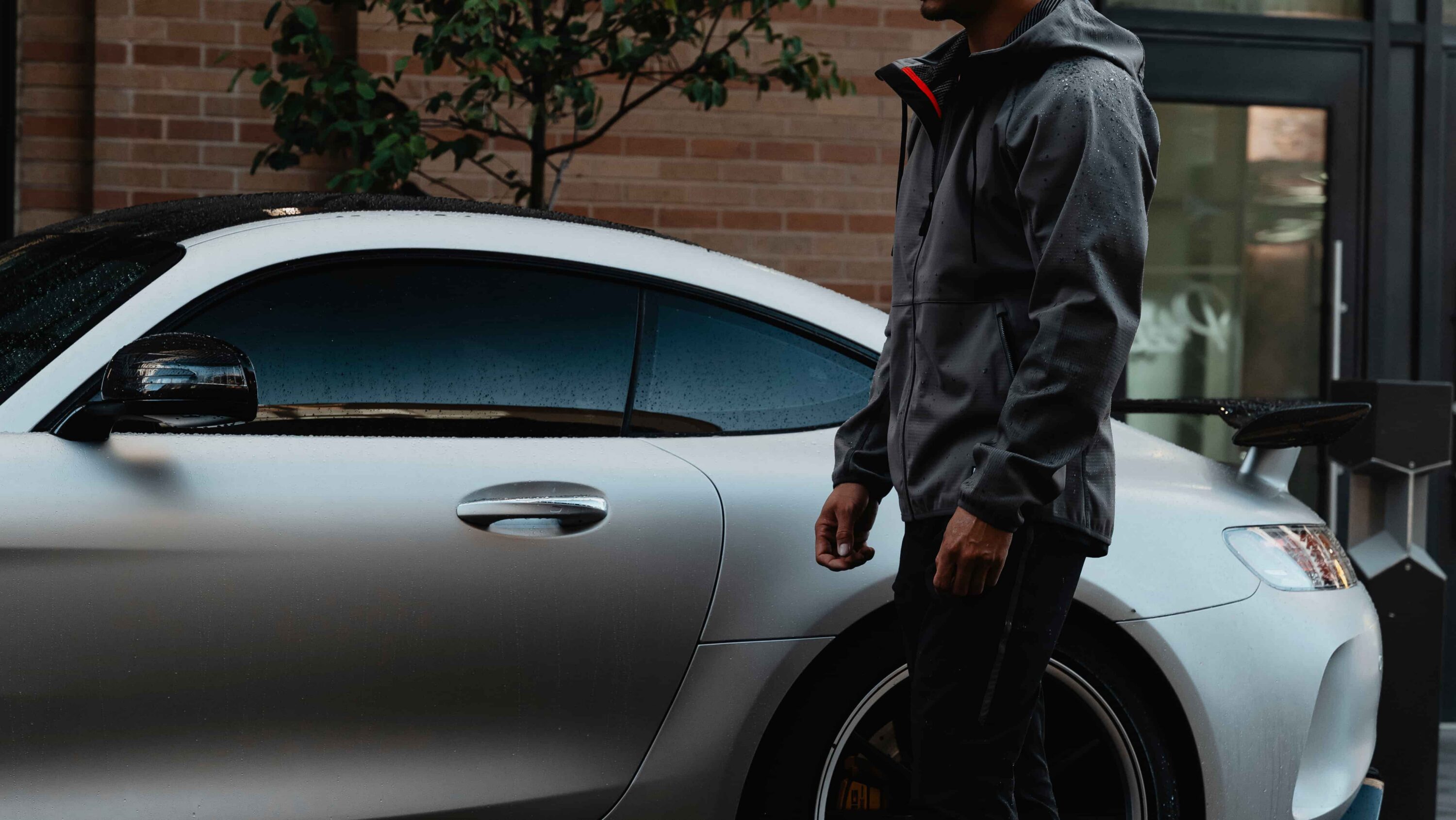 Man wearing the LVRY PACE water-resistant jacket standing beside a rain-wet silver sports car, showing the jacket's PFC-free double-knit construction holding up in wet conditions.