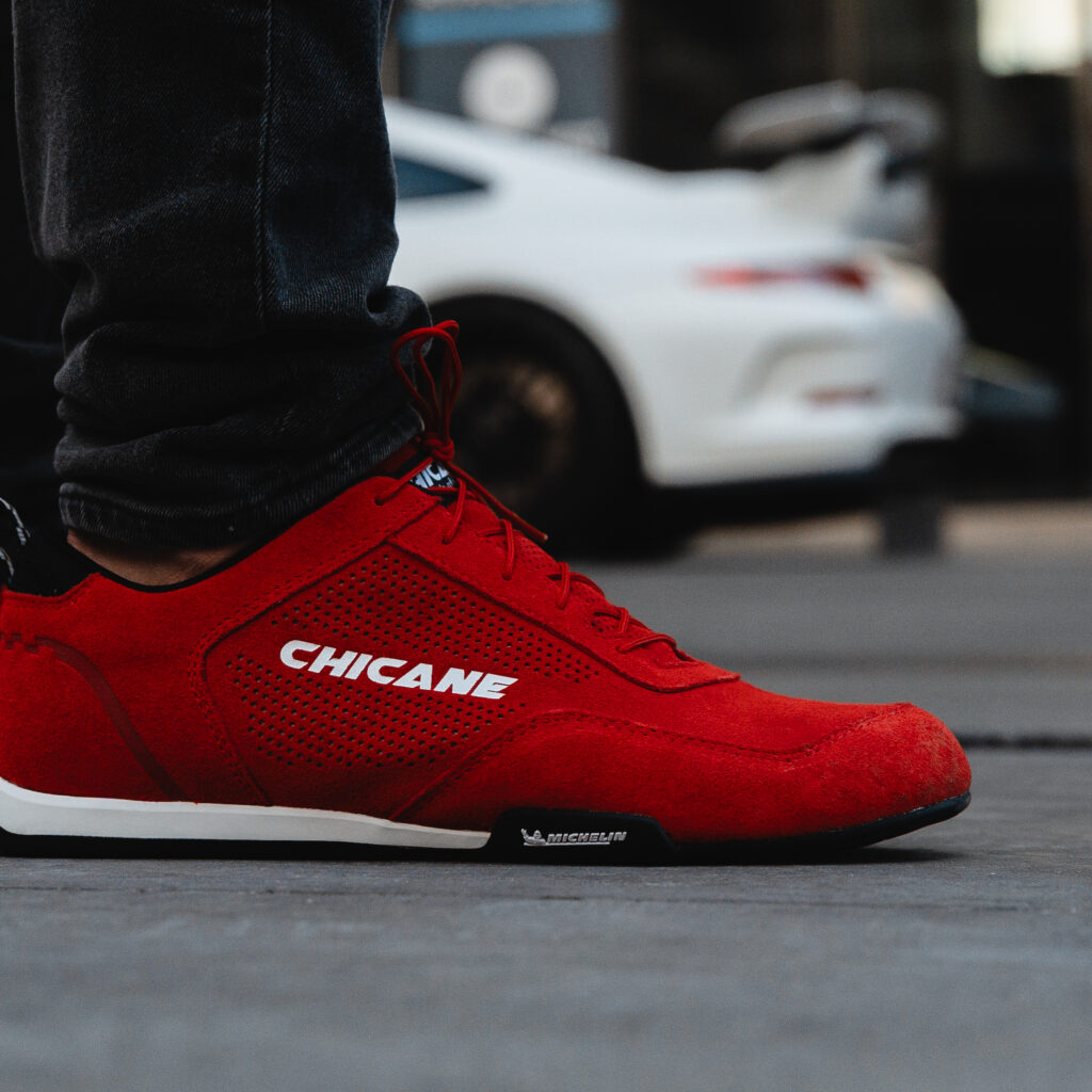 Close-up of red suede Chicane shoes with Michelin tread, worn beside a white Porsche, showing the suede upper texture covered in this motorsport footwear care guide.