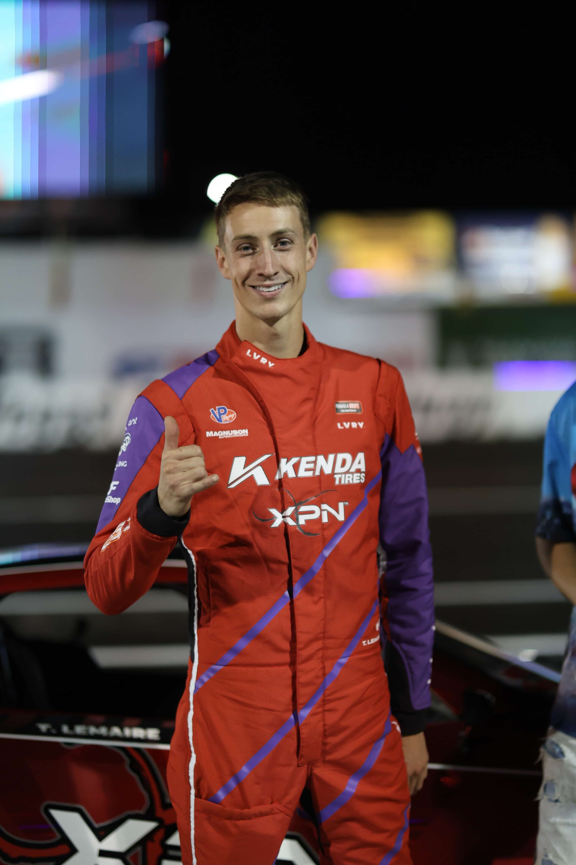 Tommy Lemaire in a red and purple Formula Drift DRVR suit with LVRY and Kenda Tires logos, giving a shaka at a night event.