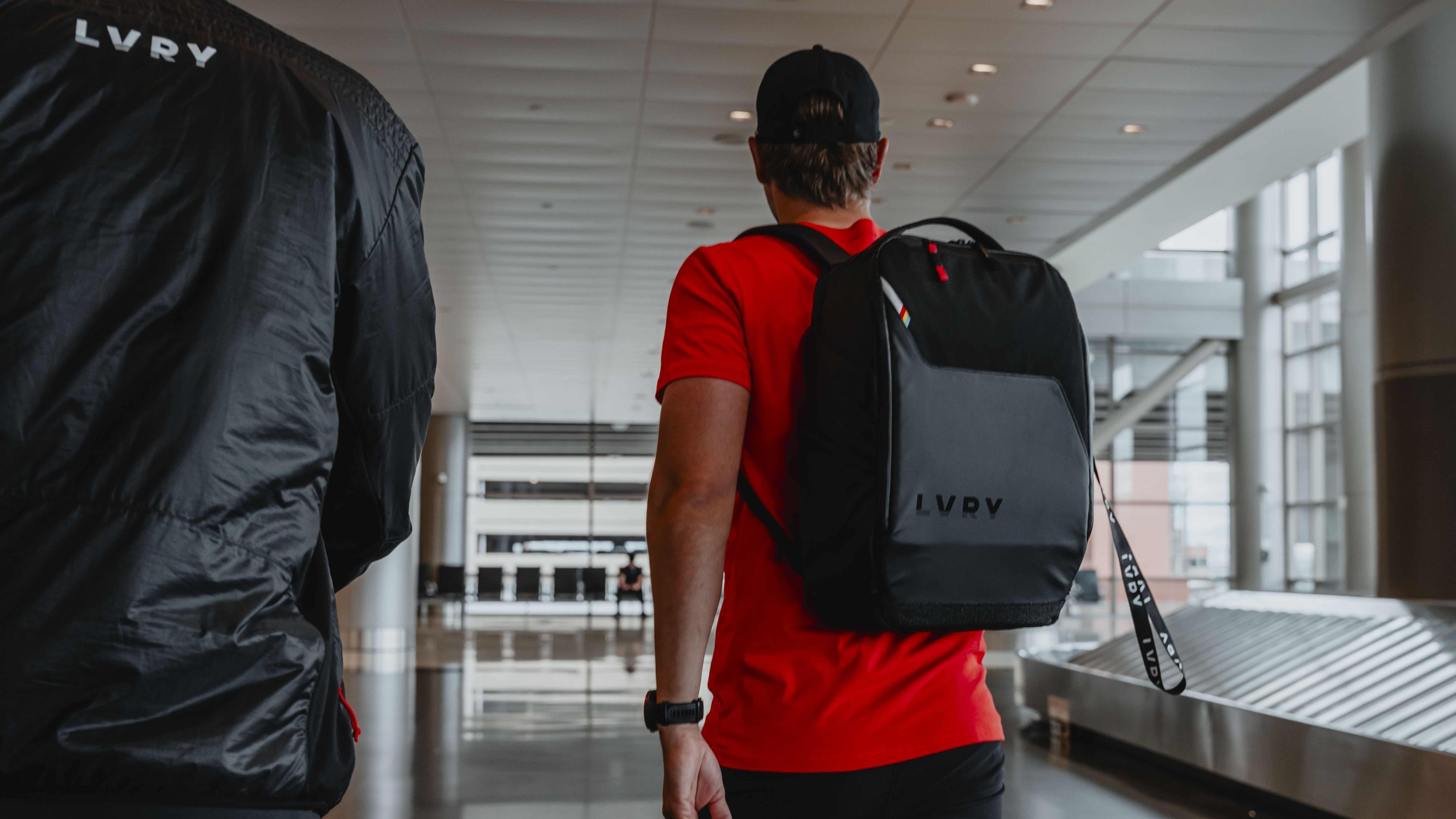 Man in a red tee walking through an airport past the baggage carousel with the LVRY PACK on his back.