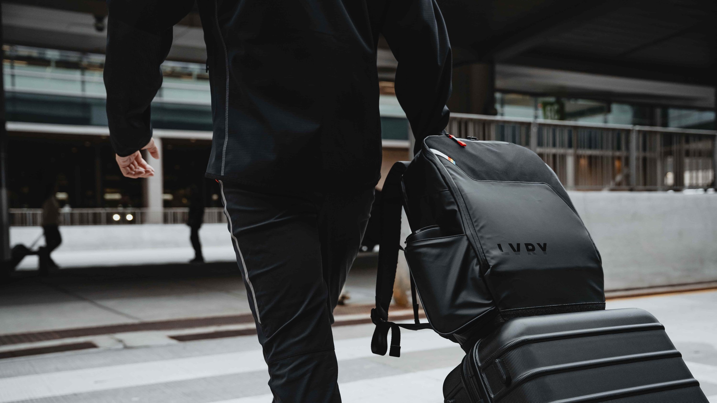 LVRY PACK riding on top of a rolling suitcase as a traveler heads across the airport crosswalk, with a bottle in the side pocket.