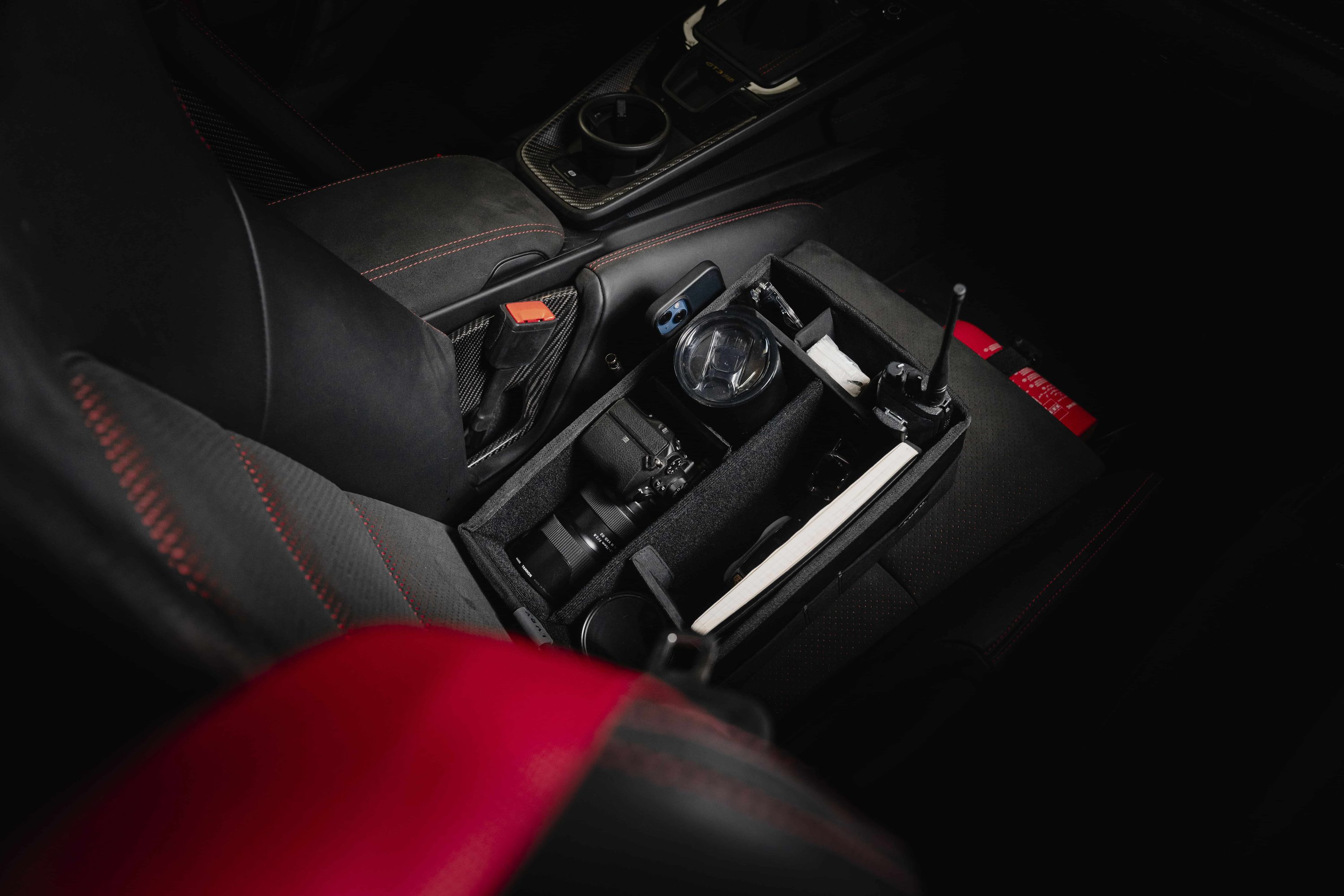 LVRY CO-DRVR organizer sitting on the passenger seat of a race car, holding a camera, radio, tumbler, phone, and notebook in padded compartments.
