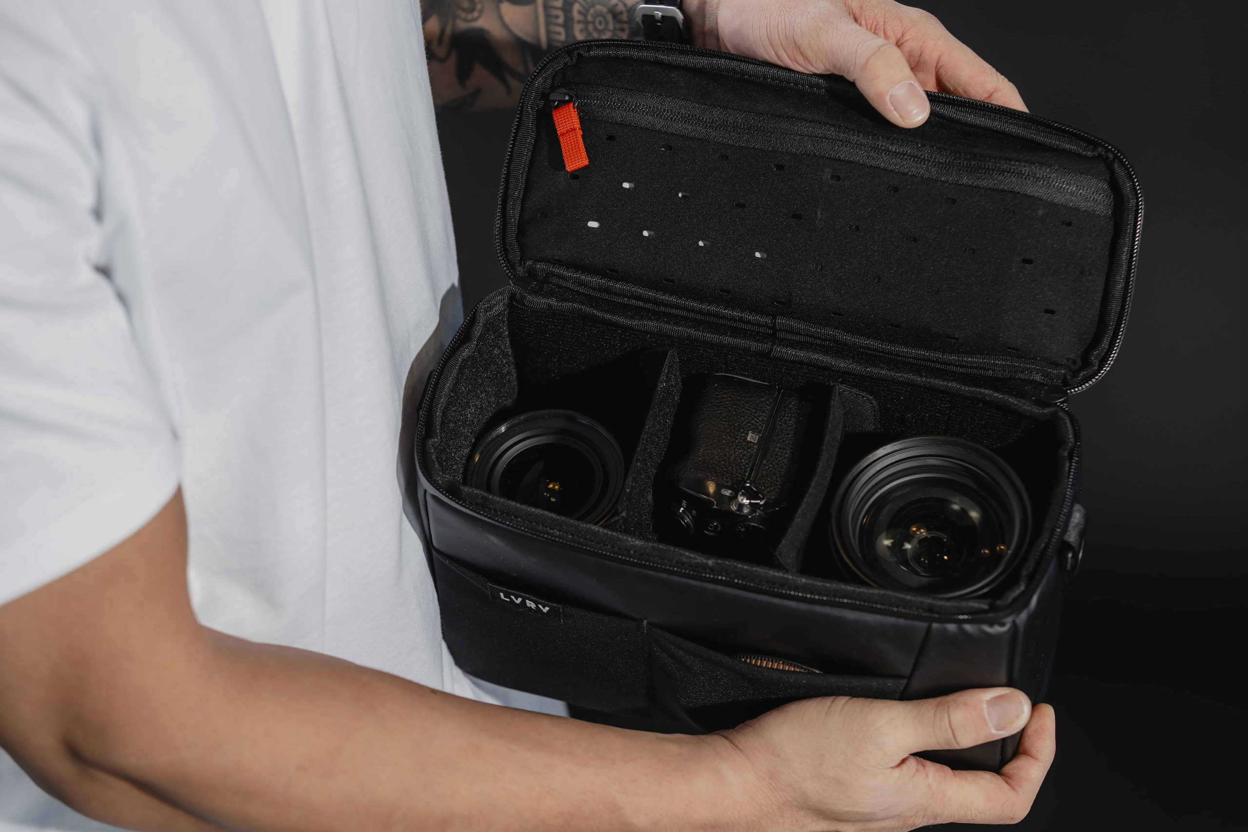 Open LVRY PITBOX with adjustable padded partitions holding a mirrorless camera and two lenses, with an orange zipper pull on the lid.