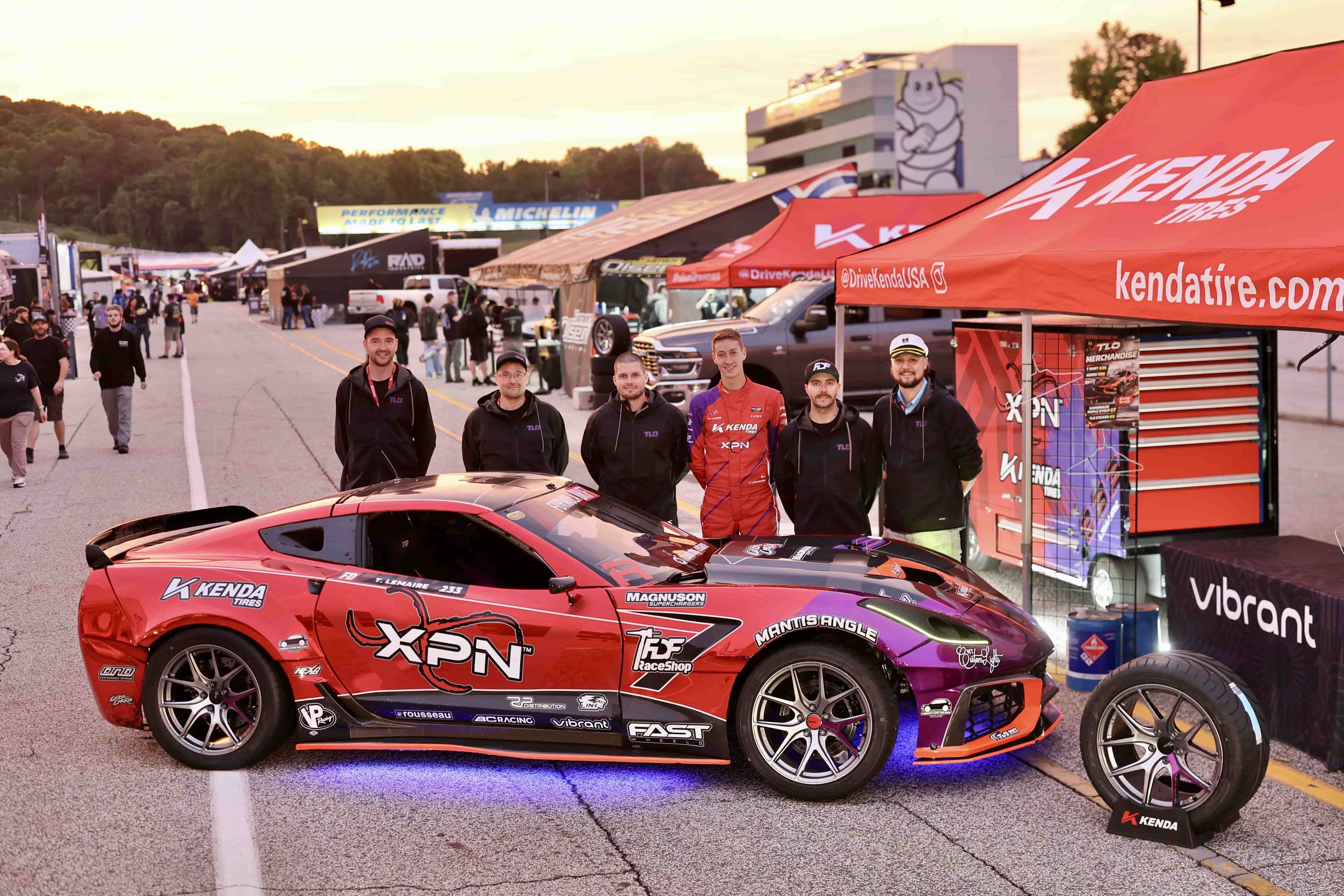 Tommy Lemaire and team posing with the red and purple Corvette C7 drift car and matching pit cart at Road Atlanta at sunset.